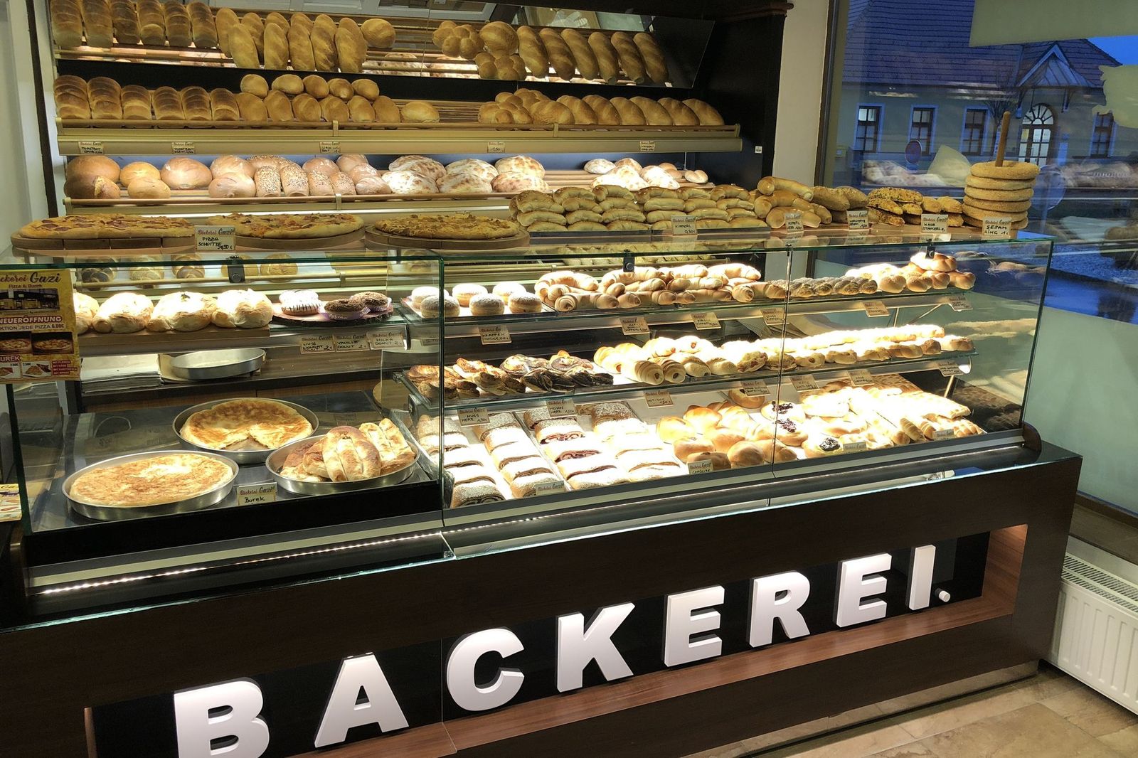 Verkaufstheke der Bäckerei Gazi: Regale mit Brot und Gebäck, darunter eine Vitrine mit Pizza, Burek und Gebäck, an der Theke der Schriftzug „Bäckerei“